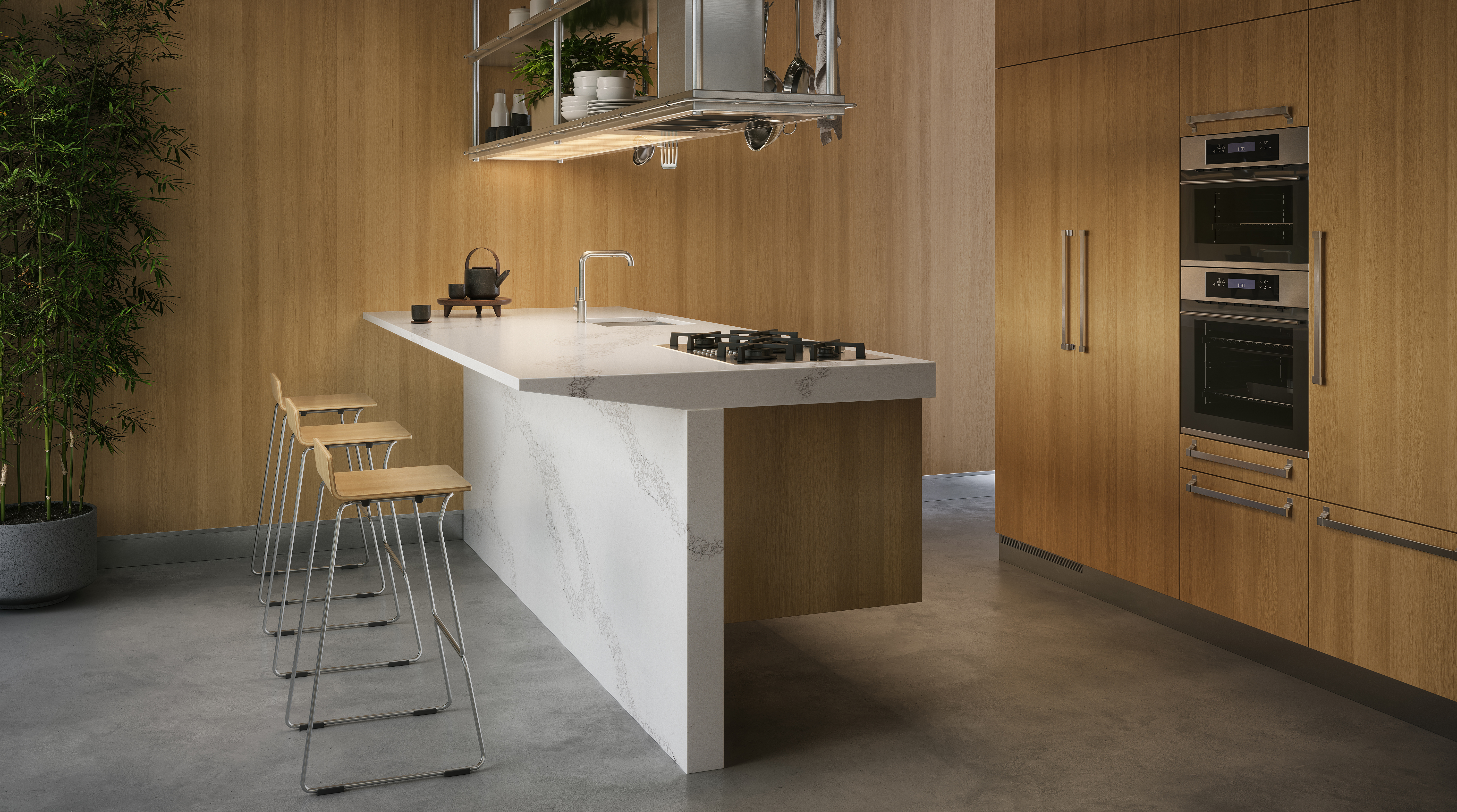 Modern kitchen with wooden cabinets, white island, built-in ovens, bar stools, and potted plant.