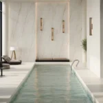 Indoor modern swimming pool with lounge chairs, plants, large windows, and Caesarstone porcelain 502 Sleet cladding walls.